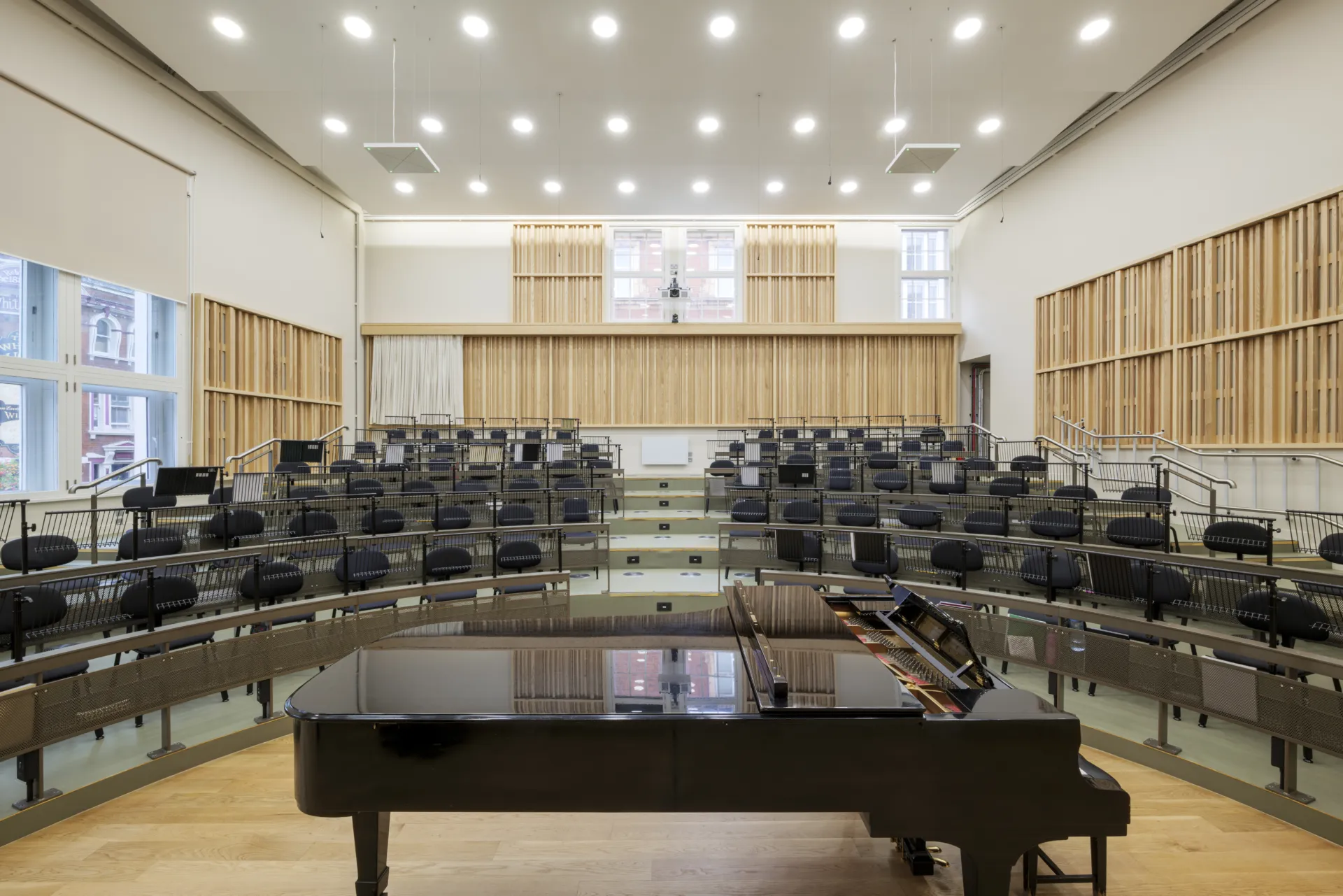 Black grand piano inside a music rehearsal room with several rows of seats and wood acoustic panelling